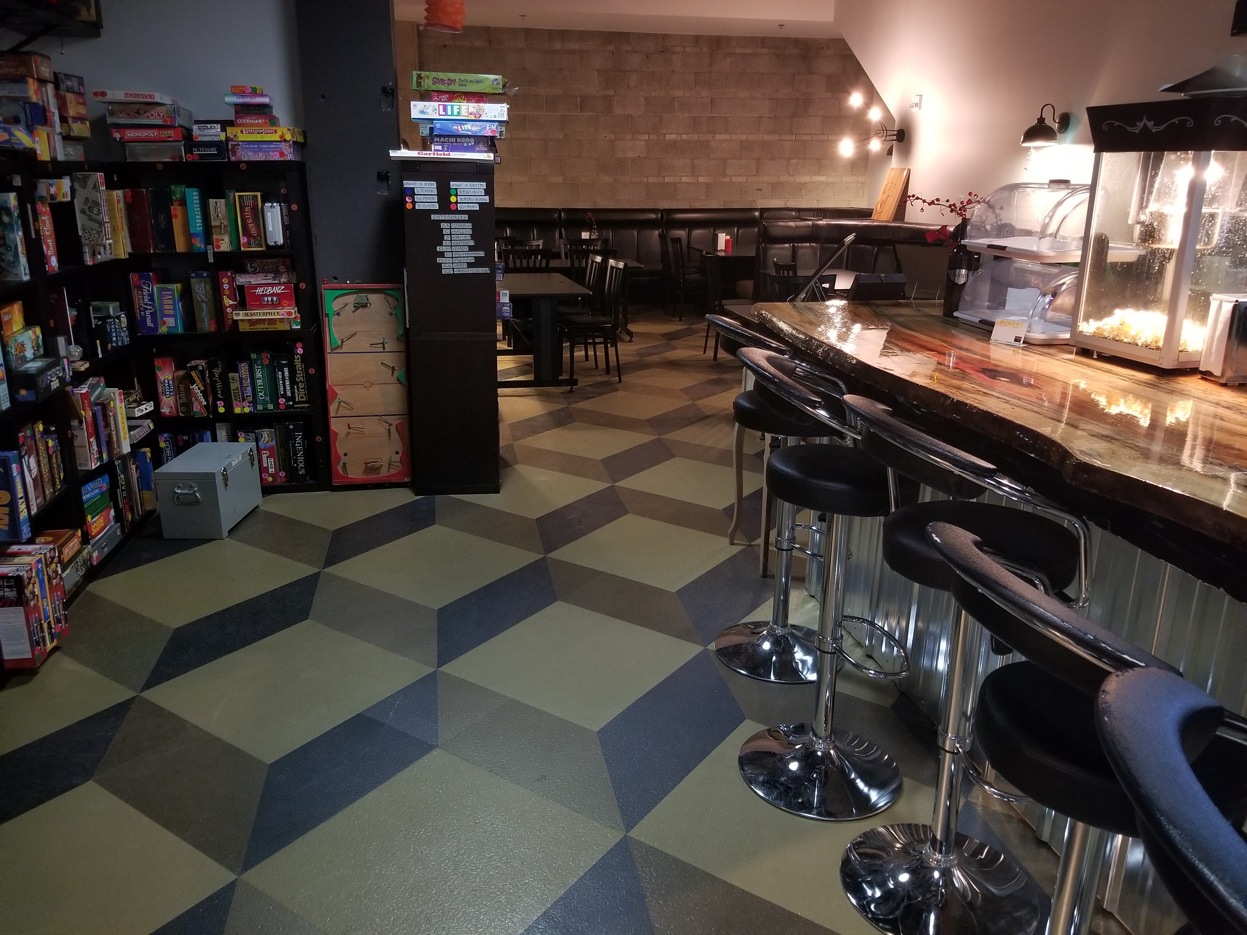 Picture of a cafe with tall chairs at a counter, shelves with board games, and at the back is a lounge with tables and chairs.